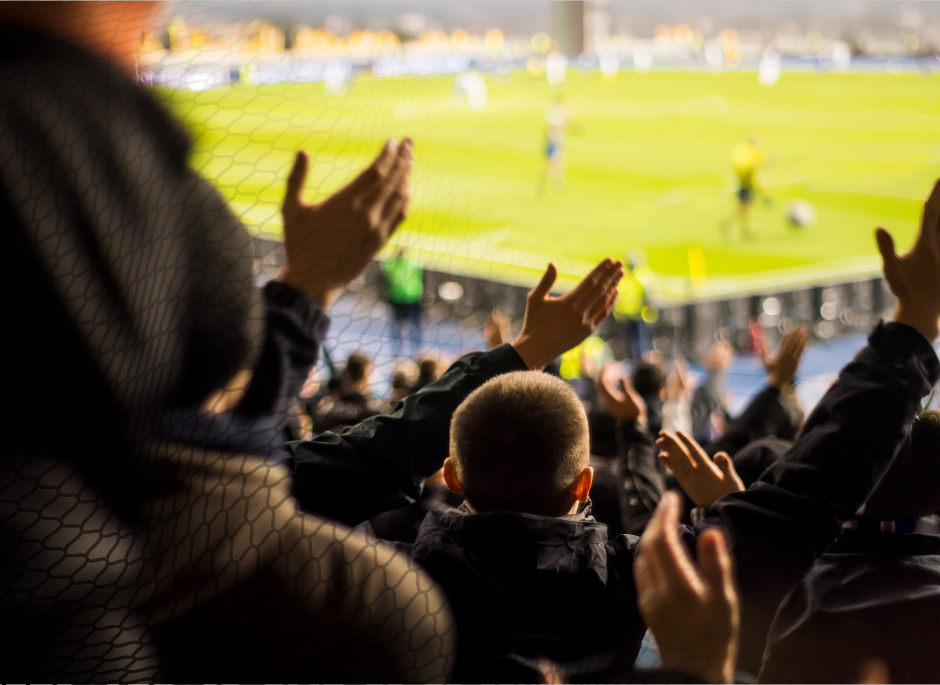 fans, including a small child, clap as a soccer match is going on in the background 
