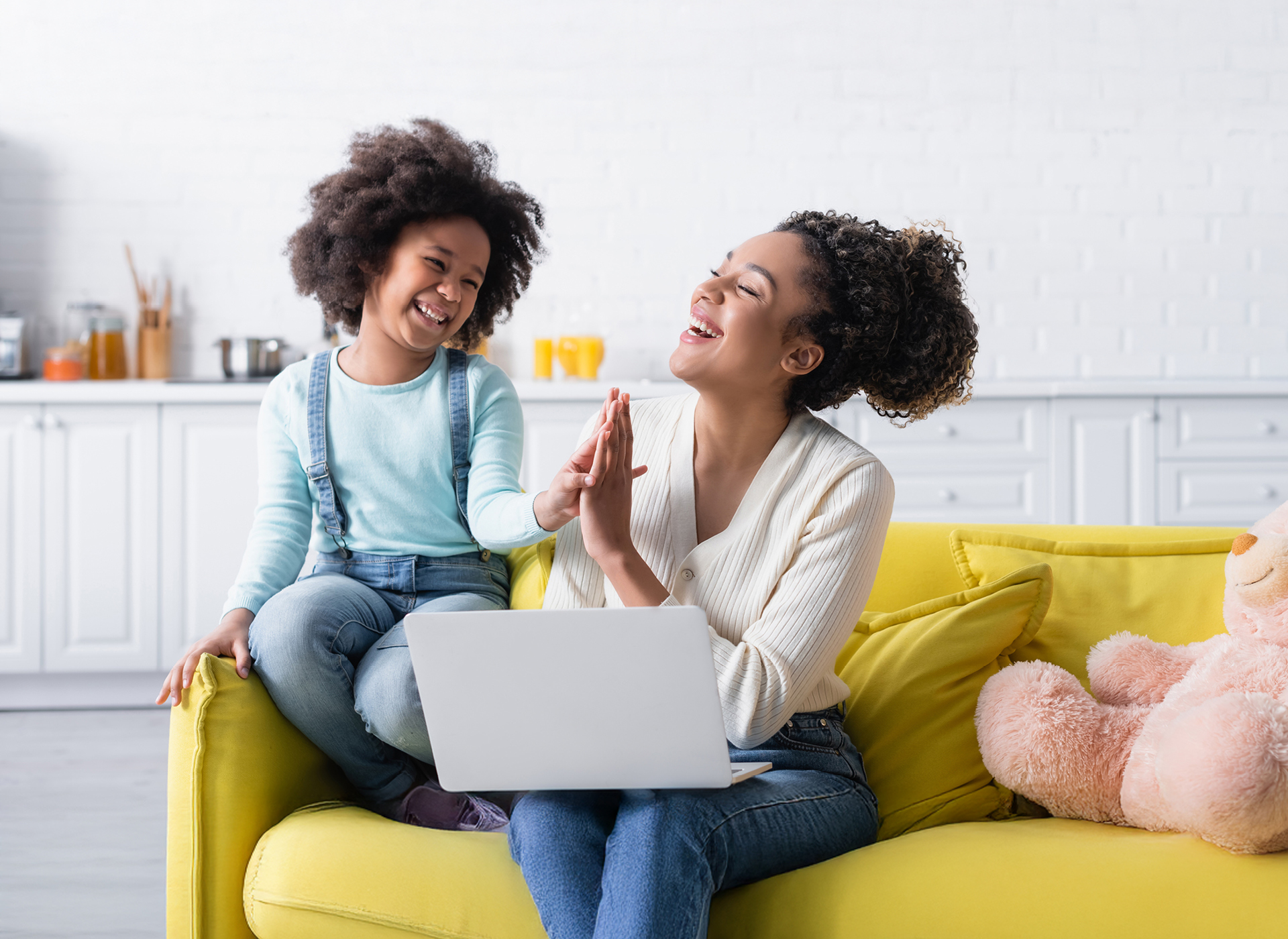 hispanic mom and young daughter sit on yellow couch with laptop out, smiling