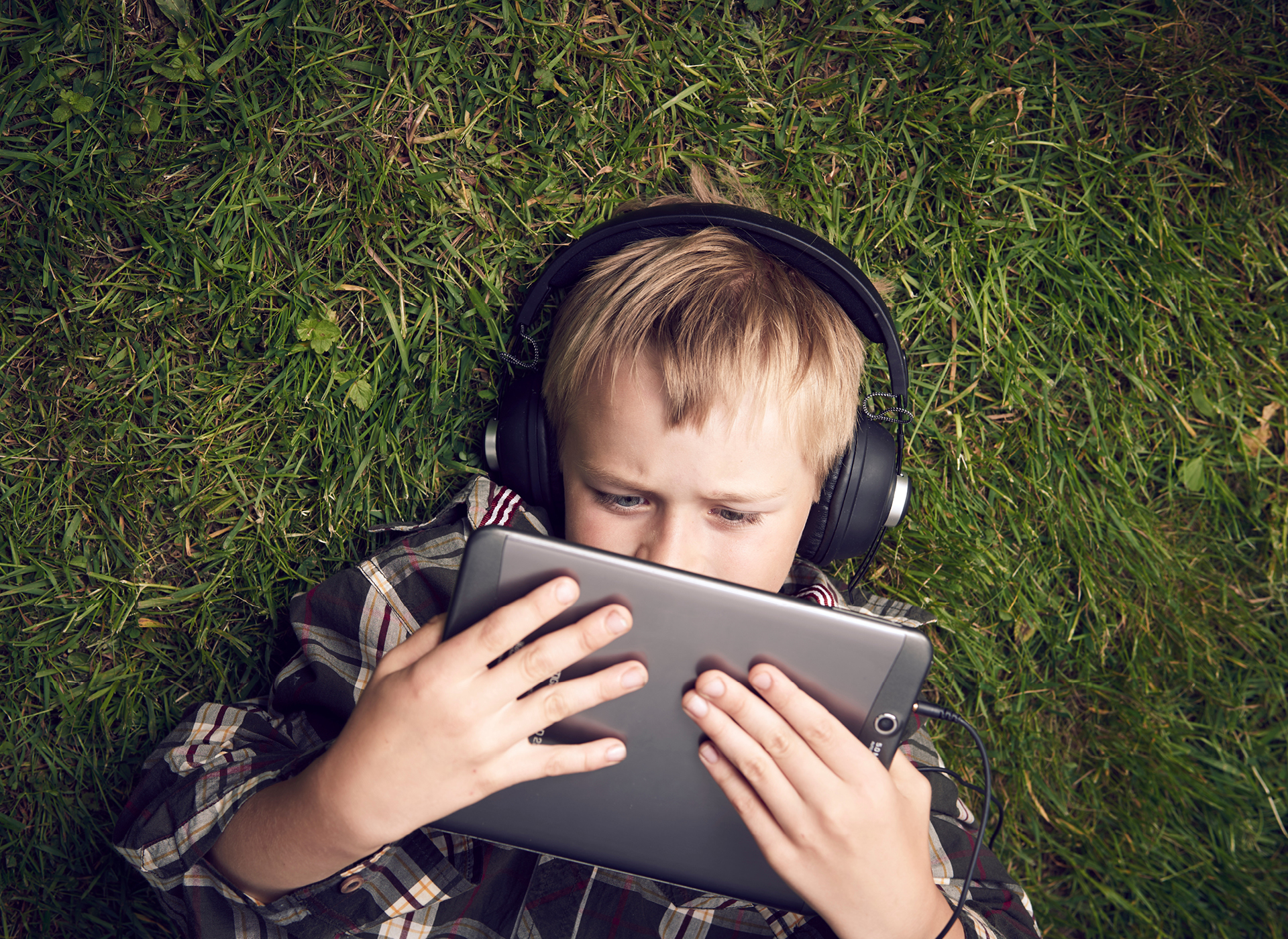 little boy lying in grass with headphones on looking at tablet